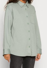 Camicia a maniche lunghe di un verde tenue, realizzata in tessuto morbido, con chiusura a bottoni sul davanti, ampio colletto e una tasca sul petto.
