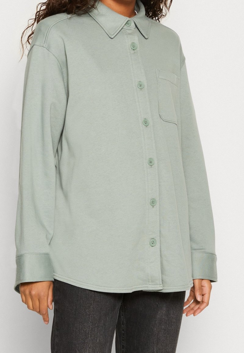 Camicia a maniche lunghe di un verde tenue, realizzata in tessuto morbido, con chiusura a bottoni sul davanti, ampio colletto e una tasca sul petto.