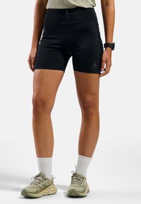 Zwarte sportshorts met een hoge tailleband, voorzien van een ritssluiting aan de voorkant en een logo-detail. Gecombineerd met olijfgroene sportschoenen en witte sokken.