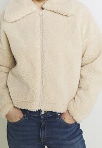 Giacca in pile beige con una superficie morbida e testurizzata, dotata di chiusura a zip frontale e un ampio colletto, abbinata a jeans in denim blu.