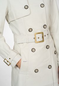 White leather trench coat featuring double-breasted design, large dark buttons, a belted waist, and stitched accents on the cuffs and pockets.