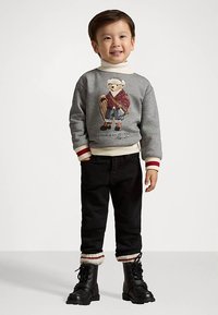 Χαμογελαστός αγόρι που φοράει ένα γκρι πουλόβερ με γραφικό αρκούδας, μαύρα παντελόνια με αναδιπλωμένα μπατζάκια με κόκκινες και λευκές ρίγες, και μαύρες υποδήσεις με κορδόνια.