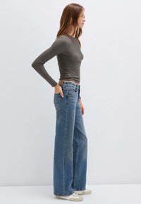 Camicia grigia a maniche lunghe aderente abbinata a jeans blu a vita alta e gamba larga. I jeans presentano un lavaggio chiaro e una piccola etichetta in pelle marrone sul retro.