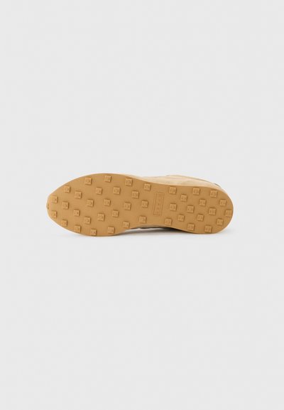 Semelle de chaussure avec une bande de roulement en caoutchouc couleur fauve, présentant des clous carrés disposés en motif de grille sur un fond clair uni.