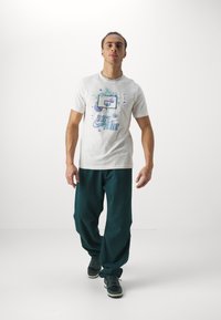 Camiseta blanca de algodón con estampado gráfico que presenta un aro de baloncesto y el texto "SOLO HAZLO", combinada con pantalones de chándal verdes holgados y zapatillas negras.