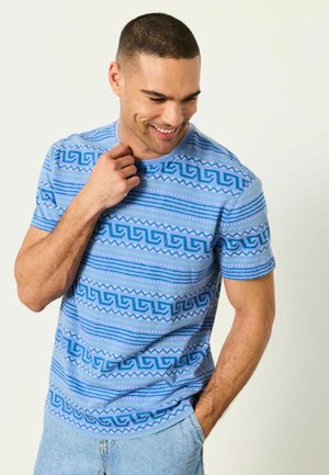 Mann lächelt mit kurzen Haaren, trägt ein blau gemustertes T-Shirt und hellblaue Jeans, eine Hand berührt den Hals, die andere in der Tasche.