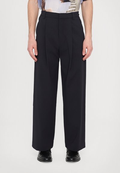 Navy tailored trousers met een brede pijp, geplooide voorkant en riemlussen, vervaardigd uit een gladde stof. Gecombineerd met gepolijste zwarte schoenen.