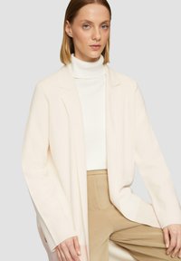 Helles rosa Cardigan aus weichem Material mit entspannter Passform, Reverskragen, offener Vorderseite und langen Ärmeln, kombiniert mit einem weißen Rollkragenpullover und beigen Hosen.