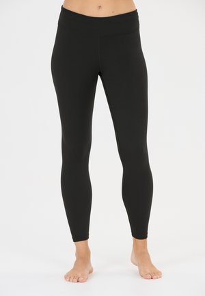 Vrouw draagt effen zwarte nauwsluitende leggings, staat op blote voeten tegen een witte achtergrond, afbeelding bij de taille en enkels bijgesneden.