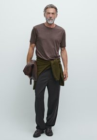 Brun bomulds T-shirt, grøn sweater bundet omkring livet, grå bukser og sorte sko. Teksturen ser blød og afslappet ud.