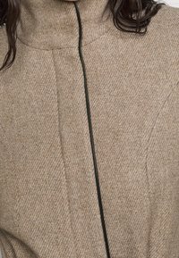 Abito in misto lana beige con colletto alto, caratterizzato da dettagli in zip nera, fine motivo houndstooth e cuciture strutturate.