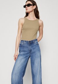 Woman wearing black rectangular sunglasses, sleeveless beige top, and loose-fitting blue jeans, standing with one hand behind back.