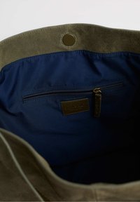 Alyvuogių žalios zomšos maišo vidus su tamsiai mėlynu audinio pamušalu. Yra užtrauktuko kišenė ir ruda prekės ženklo etiketė. Apvalus atidarymas viršuje.
