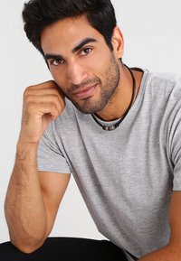 Graues T-Shirt mit kurzen Ärmeln und glatter Textur, kombiniert mit einer schwarzen Halskette mit metallischen Akzenten. Der Hintergrund ist neutral.