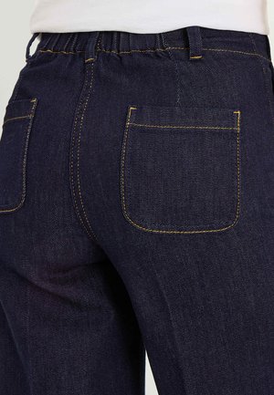 Gros plan sur un jean en denim bleu foncé avec une ceinture élastique et deux poches arrière avec des coutures jaunes, porté avec une chemise blanche.