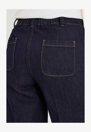 Gros plan sur un jean en denim bleu foncé avec une ceinture élastique et deux poches arrière avec des coutures jaunes, porté avec une chemise blanche.