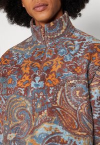 Pullover in fleece con colletto alto, caratterizzato da un motivo paisley nei toni del marrone, blu e arancio. Chiusura con zip e logo del marchio ricamato sul petto.