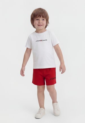 Ungt barn med lyst brunt hår iført en hvid Jordan t-shirt, røde Jordan shorts og hvide sneakers, stående på en ensfarvet baggrund.