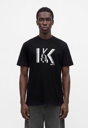 Mand står foran, iført en sort t-shirt med stort hvidt "K" og stiliseret profilgrafik foran mod en ensfarvet hvid baggrund.