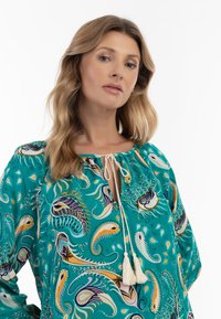 Blusa turchese con motivi paisley, caratterizzata da un colletto a chiave e lacci con pon pon. Realizzata in un tessuto leggero con un fit rilassato.