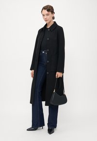 Donna che indossa un lungo cappotto nero con bottoni, jeans blu scuro, tacchi a punta neri, mentre tiene una borsa nera davanti a uno sfondo bianco semplice.