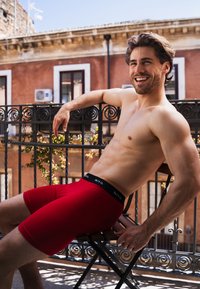 Röda boxerkalsonger med svart midjeband och vit text, sittande på en trästol mot en bakgrund av en balkong.