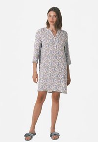 Valkoinen mekko, jossa on kukka- ja lehtikuvioita violeteissa ja sinisissä sävyissä, kolmen neljänneksen hihat ja edessä nappilista.