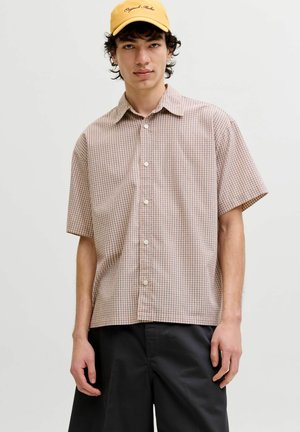 Ung mand iført en beige ternet kortærmet skjorte, sorte bukser og en gul baseballkasket med kursiv tekst.