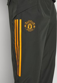Svarta träningsbyxor med en orange Manchester United-logotyp och tre vertikala orange ränder längs vänster sida. Lättviktig, texturerad tyg.