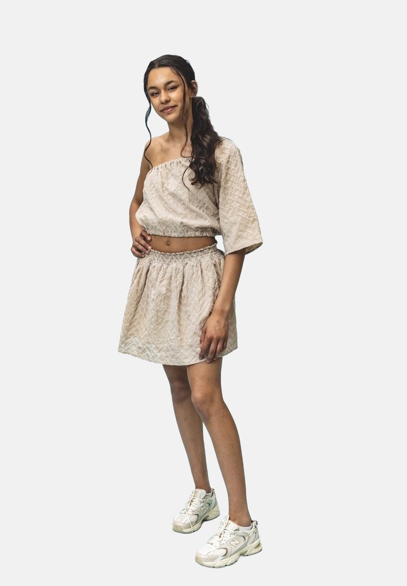 Beige twee-delige outfit met een gestructureerde off-the-shoulder top met brede mouwen en een bijpassende rok met rekbare taille, gecombineerd met witte sneakers.