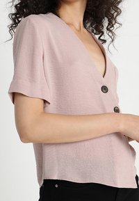 Kvinna klädd i en ljusrosa kortärmad blus med stora mörka knappar och svarta byxor, med armarna i kors vid midjan, lockigt mörkt hår synligt.