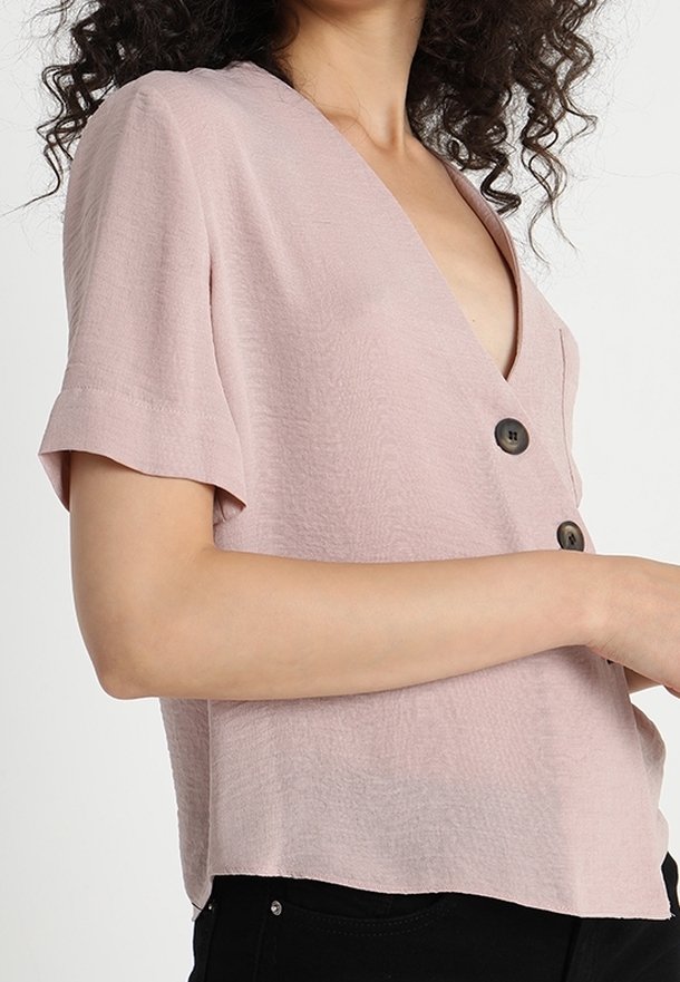 Kvinna klädd i en ljusrosa kortärmad blus med stora mörka knappar och svarta byxor, med armarna i kors vid midjan, lockigt mörkt hår synligt.