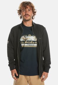 Black zip-up jacket over a blue t-shirt featuring a colorful palm and wave graphic. Smooth fabric, long sleeves, and a sporty design.