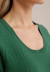 Haut en tricot texturé vert avec un large décolleté et une finition côtelée. Le tissu présente un motif à carreaux subtil.