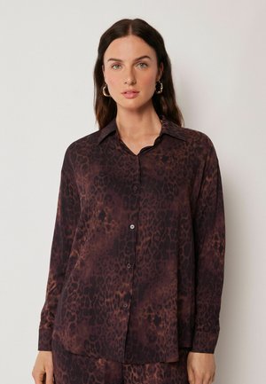 Donna con capelli lunghi e scuri che indossa orecchini a cerchio dorati e una camicia marrone scuro con stampa leopardata, davanti a uno sfondo chiaro.