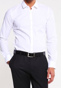 Camisa blanca de botones con cuello y mangas largas, de textura suave, acompañada de un cinturón negro con hebilla plateada, llevada con pantalones oscuros.