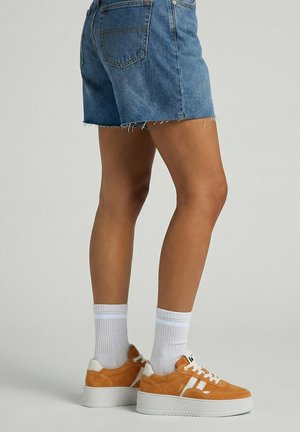 Piernas con pantalones cortos de mezclilla azul, calcetas blancas hasta el tobillo y zapatillas naranjas con cordones blancos sobre un fondo liso gris claro.