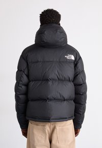 Svart puffjacka med hög krage och huva, med en studsande, vadderad design och en vit logotyp på den övre vänstra ryggen.