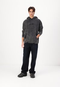 Kapucnis pulóver sötétszürkében, kopott textúrával, elülső zsebbel, sötét farmernadrággal és mintás dizájnnal rendelkező sportcipőkkel párosítva.