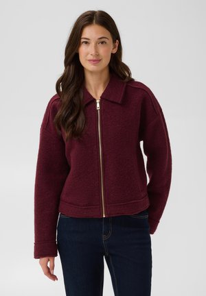 Mujer con cabello largo y oscuro, vestida con una chaqueta burdeos con cremallera y jeans azul oscuro, de pie frente a un fondo claro y liso.