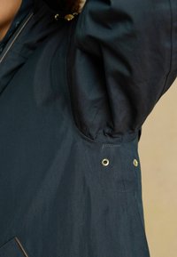 Tmavomodrá bunda z hladkej tkaniny s zlatými kovovými detailmi a pevne priliehajúcim rukávom. Viditeľné švy a očká blízko manžety.