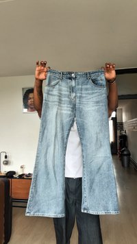 Oseba drži široke svetlo modre kavbojke pred seboj v prostoru z lesenim tlemi in pohištvom.