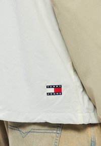 Fehér és bézs rövid ujjú ing, texturált szövettel és egy kis hímzett logóval, amely piros, fehér és kék díszítésekkel rendelkezik.