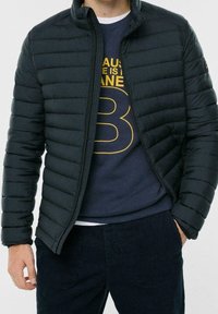 Černá prošívaná bunda s předním zipem, horizontálním prošíváním a vysokým límcem. Nositelná přes tmavě modrou mikinu s žlutým grafickým motivem.