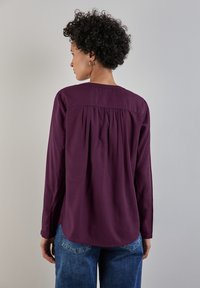 Blusa viola a maniche lunghe con dettaglio arricciato sul retro, scollo rotondo e polsini abbottonati, abbinata a jeans blu. Tessuto con texture morbida.