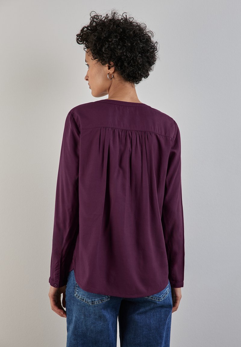 Blusa viola a maniche lunghe con dettaglio arricciato sul retro, scollo rotondo e polsini abbottonati, abbinata a jeans blu. Tessuto con texture morbida.