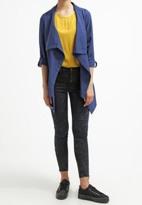 Veste drapée bleu sur un haut jaune, associée à un jean skinny noir et des baskets noires. La veste a des manches retroussées et une coupe fluide.