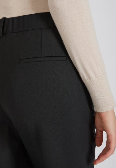 Czarne spodnie o dopasowanym kroju z boczną kieszenią i gładkim, strukturalnym materiałem. Pokazana ręka lekko spoczywa na tkaninie.