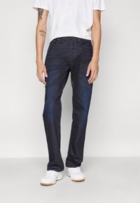 Mørkeblå denim jeans med diskret slidmærker, lige bendesign, fem lommer og orange syninger. Sammen med hvide sneakers.