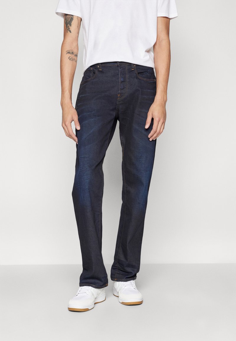 Mørkeblå denim jeans med diskret slidmærker, lige bendesign, fem lommer og orange syninger. Sammen med hvide sneakers.